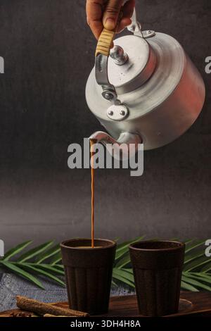 Una persona sta versando il tè masala nella tazza Bio chai. Le tazze sono su un tavolo di legno. Tagliare il chai che viene versato dal bollitore del tè. Foto Stock