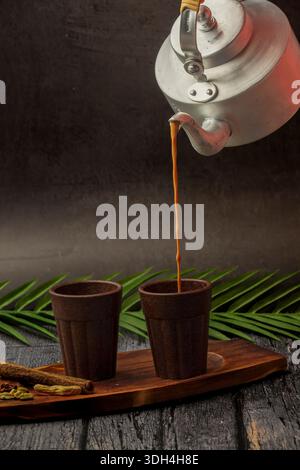 Una persona sta versando il tè masala nella tazza Bio chai. Le tazze sono su un tavolo di legno. Tagliare il chai che viene versato dal bollitore del tè. Foto Stock