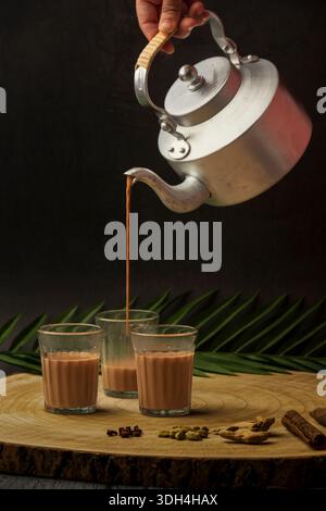 Una persona sta versando tè masala in tre tazze chai di vetro. Le tazze sono su un tavolo di legno. Tagliare il chai che viene versato dal bollitore del tè. Foto Stock