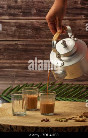 Una persona sta versando tè masala in tre tazze chai di vetro. Le tazze sono su un tavolo di legno. Tagliare il chai che viene versato dal bollitore del tè. Foto Stock