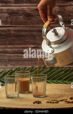 Una persona sta versando tè masala in tre tazze chai di vetro. Le tazze sono su un tavolo di legno. Tagliare il chai che viene versato dal bollitore del tè. Foto Stock