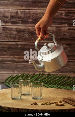 Una persona sta versando tè masala in tre tazze chai di vetro. Le tazze sono su un tavolo di legno. Tagliare il chai che viene versato dal bollitore del tè. Foto Stock