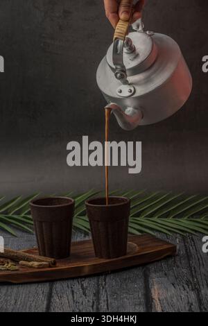 Una persona sta versando il tè masala nella tazza Bio chai. Le tazze sono su un tavolo di legno. Tagliare il chai che viene versato dal bollitore del tè. Foto Stock