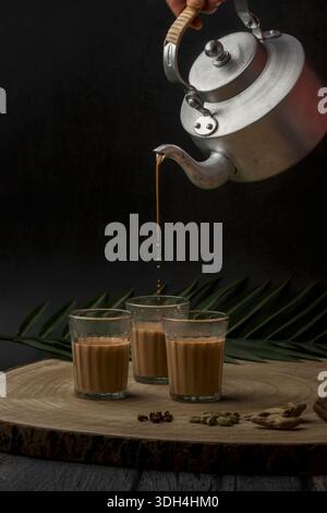 Una persona sta versando tè masala in tre tazze chai di vetro. Le tazze sono su un tavolo di legno. Tagliare il chai che viene versato dal bollitore del tè. Foto Stock