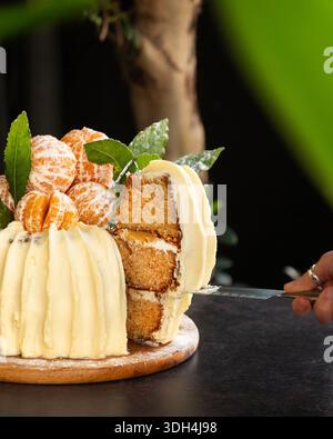 Una torta di spugne festosa decorata con mandarini interi freschi e foglie verdi, con spessa glassa di crema bianca. Foto Stock