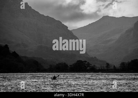 Kayak solitario che attraversa Llyn Padarn sotto ripidi pendii di montagna in bianco e nero Foto Stock