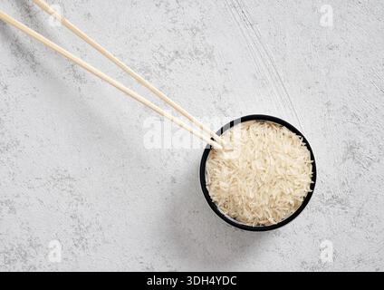 Ciotola di riso basmati crudo a grani lunghi con bacchette di legno su fondo bianco in cemento. Posate piatte, minime e naturali per cucinare Foto Stock