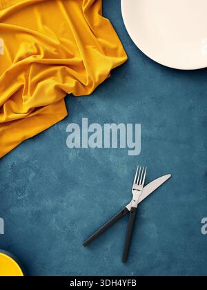 Tavolo con piatto bianco, forchetta, coltello e tovagliolo giallo su sfondo blu, per creare una base piatta minimalista per il mockup degli alimenti Foto Stock
