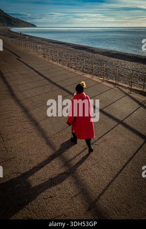 Donna con il cappotto rosso che cammina sulla spiaggia di Seaton, Devon Foto Stock