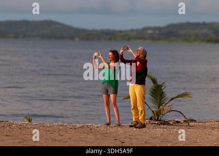 Due donne fotografano la costa con smartphone a Nadi, Fiji, in piedi su una spiaggia sabbiosa mentre la luce soffusa della sera si riflette sull'acqua. Foto Stock