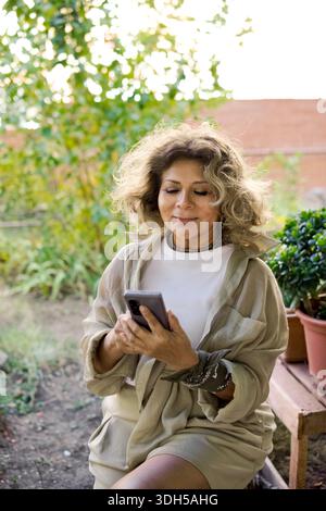 Una donna anziana serena si rilassa nel suo lussureggiante giardino, godendosi una giornata tranquilla. Sorride e si impegna con il suo smartphone, circondato da piante vivaci Foto Stock