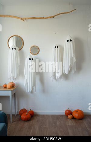 Le decorazioni bianche a fantasma sono appese su una parete con accenti in legno e specchi rotondi, circondate da zucche arancioni su un pavimento in legno, a creare una H minimalista Foto Stock