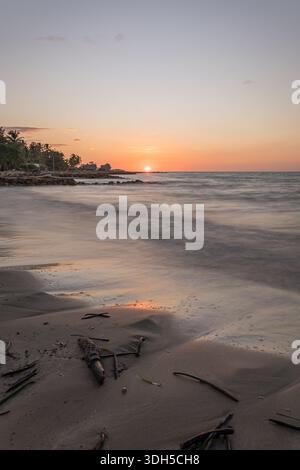 Un tramonto mozzafiato proietta tonalità vibranti su una tranquilla spiaggia caraibica. Onde dolci si infrangono sulla riva, con palme che si stagliano contro il wa Foto Stock