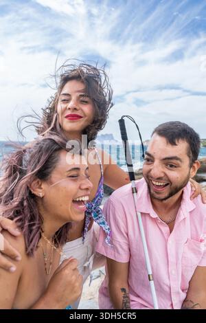 Un gruppo di amici, tra cui un individuo cieco, condividono momenti di gioia in riva al mare. I loro sorrisi riflettono l'inclusività, l'amicizia e la bellezza del div Foto Stock