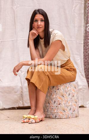 Una donna con lunghi capelli marroni dritti si siede con cura su uno sgabello a motivi geometrici in interni. Indossa una blusa leggera e pantaloni di senape, con scarpe colorate Foto Stock