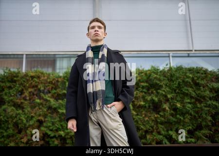 Un giovane uomo con un elegante cappotto nero e sciarpa a scacchi si erge con sicurezza su uno sfondo urbano. Le vibrazioni autunnali mettono in risalto la sua vita moderna in città Foto Stock