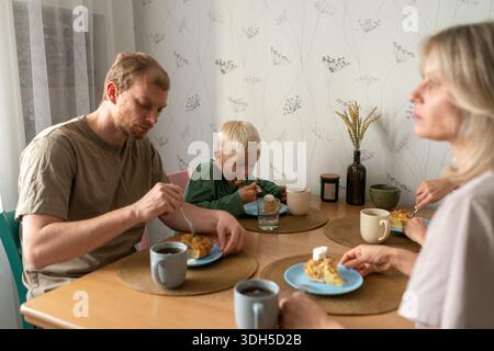 Una famiglia seduta intorno a un tavolo che gusta una colazione, creando un'atmosfera calda e accogliente con piatti, tazze e luce che scorre attraverso il vento Foto Stock