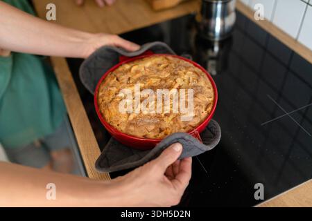 Una persona rimuove con cautela una torta di mele appena sfornata dal piano cottura utilizzando un guanto da forno. La crosta dorata e la cannella aggiungono una deliziosa e casalinga Foto Stock