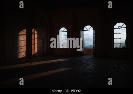 Una vista panoramica dall'interno di uno storico edificio italiano. La luce del sole proietta ombre attraverso le finestre ad arco, rivelando un pittoresco paesaggio urbano all'esterno. Foto Stock