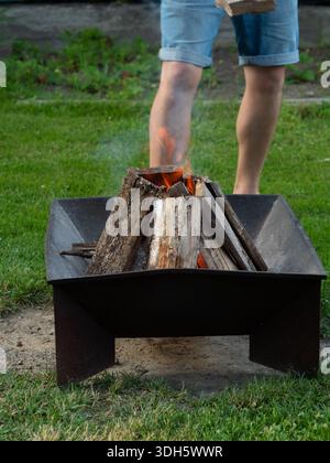 Metal Fire Pit con fiamme nel Green Backyard con le gambe dell'uomo sullo sfondo Foto Stock