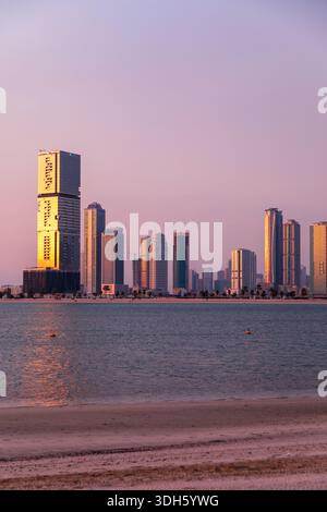 Un tranquillo paesaggio costiero di Sharjah al crepuscolo presenta grattacieli scintillanti lungo una costa tranquilla. Il caldo cielo rosa e viola si riflette sull'acqua, creato Foto Stock