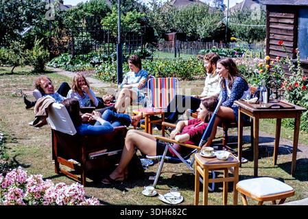 Autentico gruppo di 1970 amici che si godono una soleggiata giornata estiva nel giardino posteriore di una zona residenziale dell'Essex, Regno Unito. Ampia scelta di mobili interni, vassoi, tazze e piattini all'aperto con sedie da giardino. Moda e stili anni Settanta Foto Stock