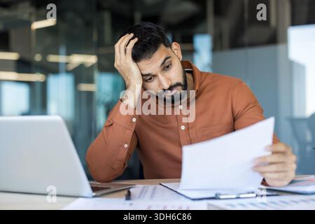 Un giovane dipendente di sesso maschile si sente stanco e frustrato mentre guarda i documenti finanziari alla sua scrivania, affrontando scadenze, stress e esaurimento professionale Foto Stock