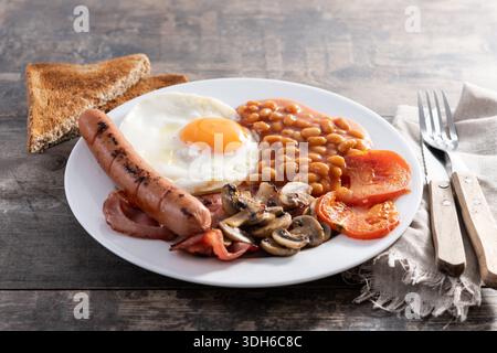 Colazione all'inglese completa con uova fritte, pancetta, salsicce e fagioli su un tavolo di legno Foto Stock