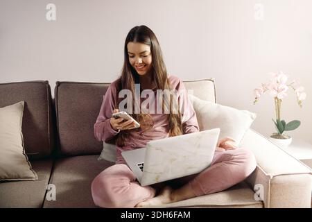 Una giovane donna siede su un divano a casa sua. Sta utilizzando un laptop in grembo e ha in mano uno smartphone, sembrando concentrata e impegnata nelle sue attività. Foto Stock