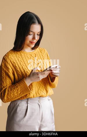 Una donna sta in piedi mentre guarda il suo smartphone con un leggero sorriso. Indossa un maglione giallo e pantaloni di colore chiaro, con sfondo neutro Foto Stock