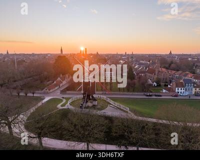 Vista aerea di un rustico mulino a vento che si erge con orgoglio contro il soffice bagliore del sole che tramonta, la sua silhouette contrasta con il paesaggio urbano di Bruges, FLA Foto Stock