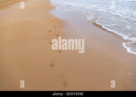 Impronte impresse nella sabbia dorata bagnata che corre parallela alla morbida schiuma marina su una spiaggia tranquilla. Foto Stock