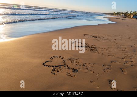 Tre cuori disegnati nella sabbia su una spiaggia dorata al tramonto, con impronte e onde oceaniche sullo sfondo. Foto Stock