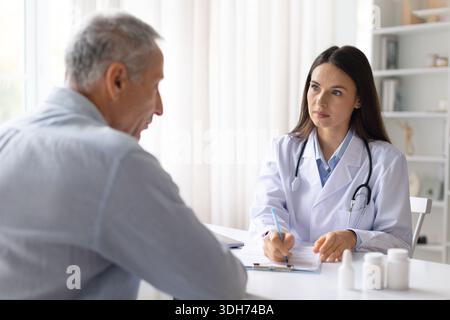 Donna medico consulenza paziente di sesso maschile senior alla scrivania e scrittura note mediche Foto Stock