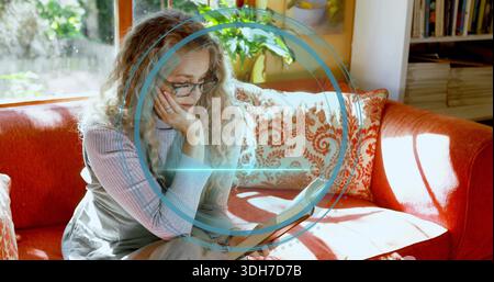 Donna anziana seduta che legge un tablet a casa su un divano rosso con specifiche, parte superiore a coste, rivestimento blu Foto Stock