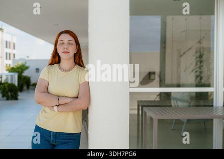 Giovane donna dai capelli rossi appoggiata a una colonna con le braccia incrociate sulla terrazza di un appartamento, ritratto urbano sicuro. Foto Stock