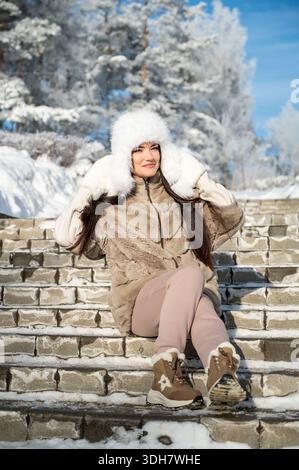 Donna che indossa un cappello di pelliccia, un cappotto caldo e guanti seduti su scale di pietra coperte di neve nel parco invernale. Atmosfera accogliente, giornate fredde e soleggiate, moda e relax stagionali Foto Stock