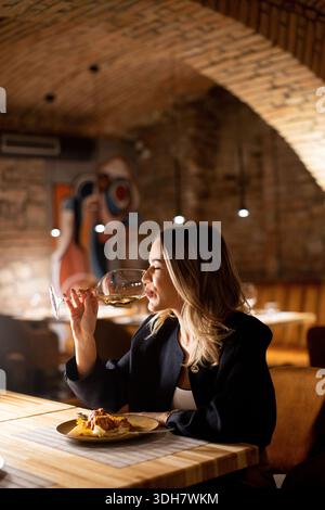 Una donna siede ad un tavolo di legno in un ristorante. Tiene un bicchiere di vino e ha un aspetto premuroso. L'ambiente presenta pareti in mattoni e illuminazione soffusa. IT Foto Stock