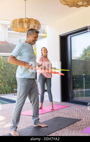Coppia senior che pratica esercizio leggero sul patio coperto a bordo piscina su tappetini per yoga, tenendo anelli Foto Stock