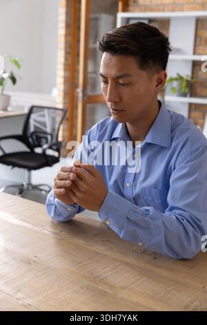 Uomo asiatico seduto a un tavolo di legno in ufficio, indossa camicia azzurra e stringe le mani Foto Stock
