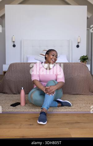 Donna afro-americana seduta su un tappeto intrecciato accanto al letto, con la parte superiore rosa, leggings color tè, con il telefono in mano Foto Stock