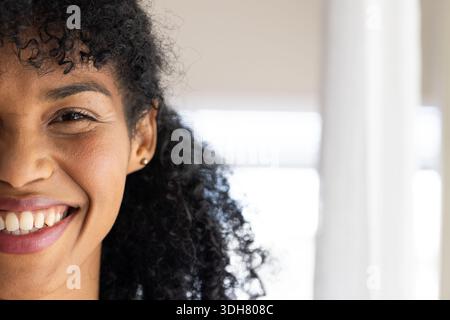 Donna afroamericana sorridente e davanti alla macchina fotografica da una finestra luminosa che mostra persiane e orecchini con borchie dorate Foto Stock