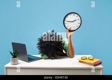 Studente maschio nero stanco sdraiato alla scrivania con computer portatile e libri, tenendo l'orologio e mettendo la testa a portata di mano, sfondo blu Foto Stock