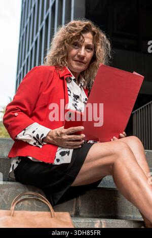 Donna d'affari che controlla lo smartphone tenendo la cartella rossa sulle scale Foto Stock