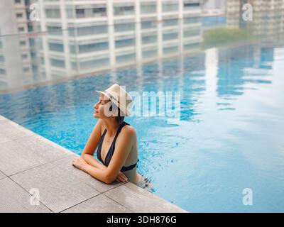 Giovane donna asiatica che si rilassa a bordo piscina all'hotel Kuala Lumpur con vista sui grattacieli circostanti, godendo del tempo libero in un vivace ambiente urbano. Foto Stock