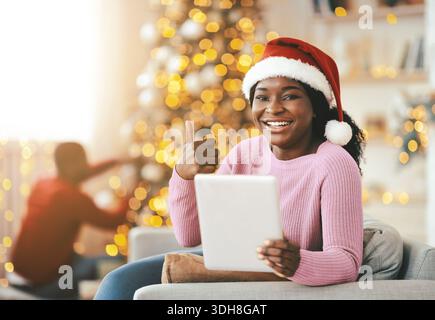 Una donna sorridente che indossa un cappello di Babbo Natale si siede sul divano che regge un tablet nel salotto delle feste Foto Stock