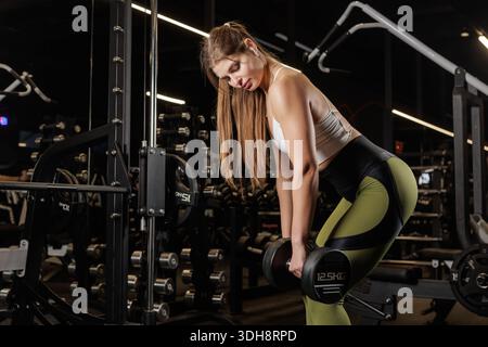 Focused Woman esegue una fila di manubri con postura a cerniera all'anca in una moderna palestra per la forza della schiena e la motivazione per il fitness Foto Stock