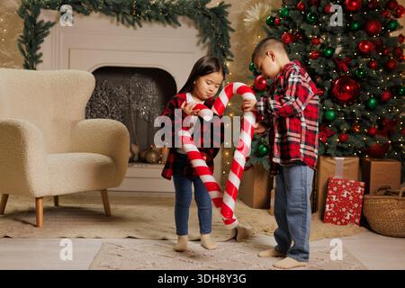 Felici bambini asiatici che giocano con grandi dolci caramelle lecca-lecca in una camera accogliente decorata con alberi di natale e ghirlande, buon natale Foto Stock