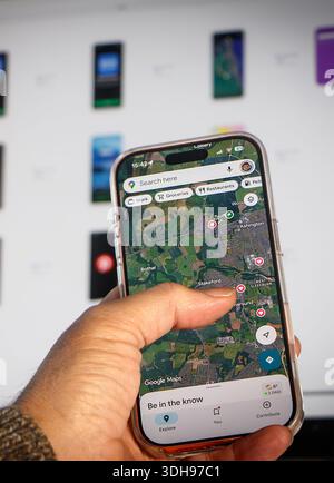 Persona che tiene in mano lo smartphone utilizzando l'app di navigazione Google Maps mentre gli schermi digitali sono visibili sfocati sullo sfondo Foto Stock