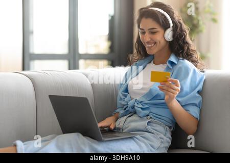 Shopping online. Felice signora caucasica con computer portatile e carta di credito seduta sul divano a casa, acquisto di merci in negozio web Foto Stock